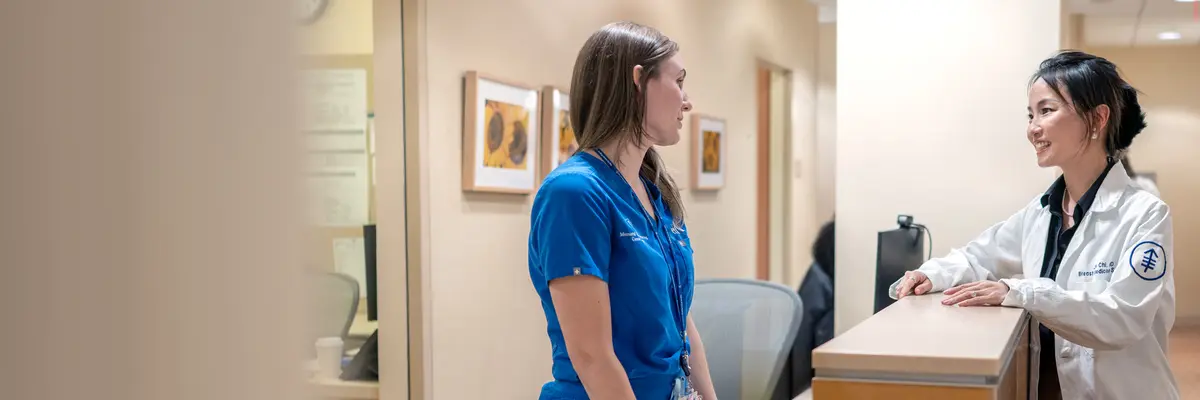 Female doctor in a white coat standing at a desk speaking with a female nurse wearing blue scrubs.