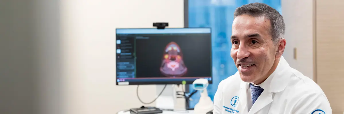 Head and neck surgeon Dr. Babak Givi, wearing a white doctors’ coat, is sitting in an MSK exam room. 