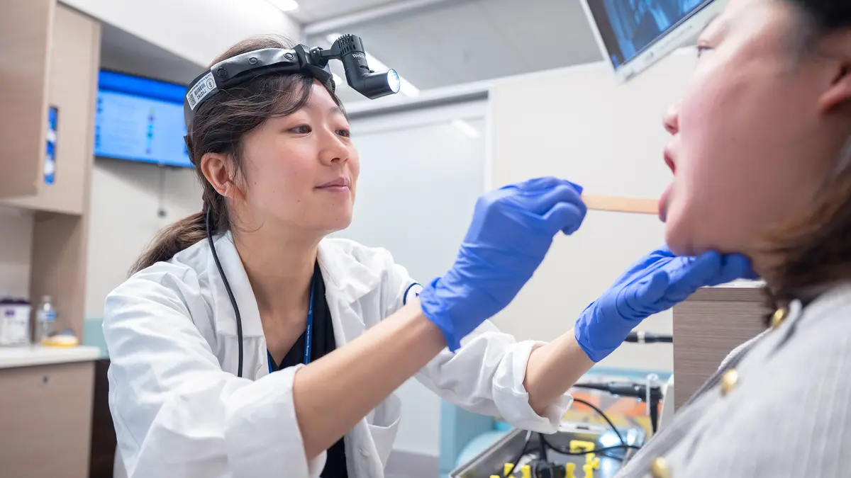 An MSK surgeon, wearing a headlamp, uses her gloved hands and a tongue depressor to examine her patient’s mouth. 