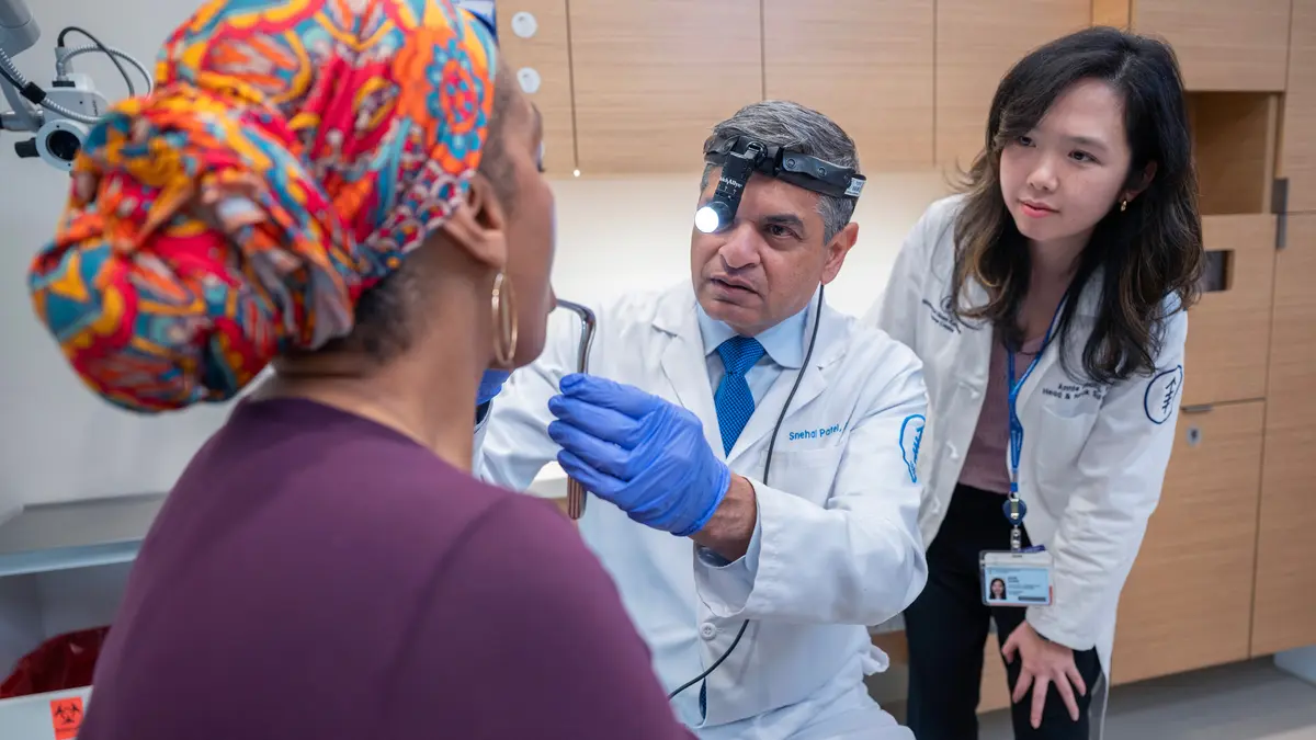 A seated MSK doctor uses a medical head lamp and gloved hands to examine his patient while another care team member stands behind him, observing. 
